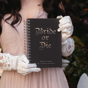 May include: A black spiral-bound wedding planner with the words "Bride or Die" in gold script. The planner also includes the text "Emmy & Gabe our wedding planner". The planner is held by a person wearing white gloves and a pink dress.