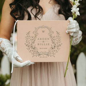May include: A blush-colored wedding guest book with a decorative floral frame and the names "Arden & Billie Wilson" printed in black. A white ribbon is attached to the spine. The date "05.10.25" is printed below the names.