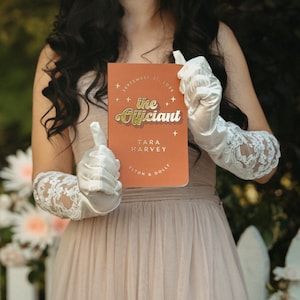 May include: An orange wedding notebook with gold lettering that reads "The Officiant" and the names "Tara Harvey, Elton & Dolly". The notebook is held by a person wearing white gloves with lace detailing.
