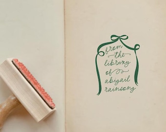 May include: An open book with aged, off-white pages and a wooden rubber stamp resting on the left page. The right page features a green ink stamp design with a ribbon border and the text, from the library of abigail rainsong, in cursive.
