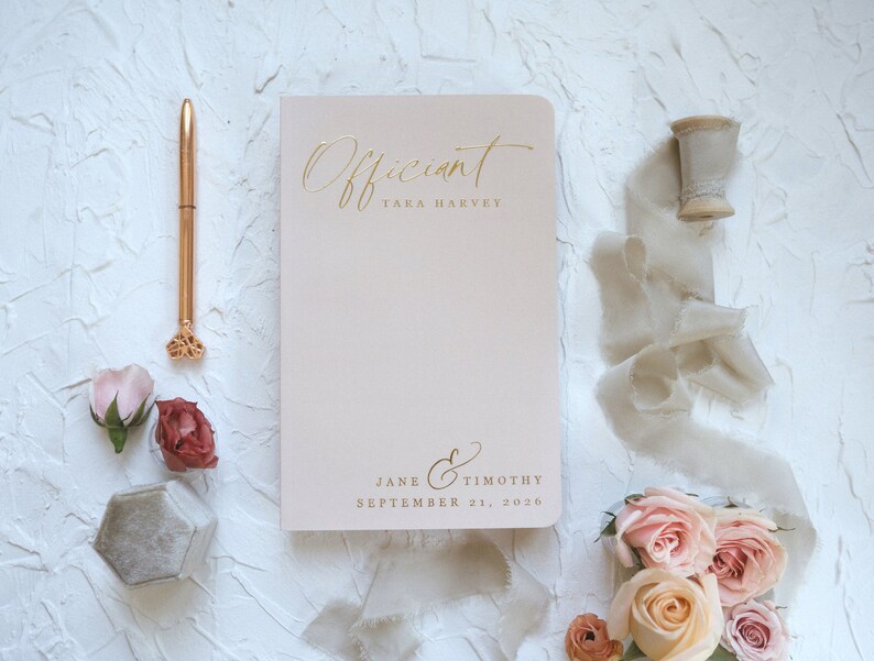 K&ouml;nnte beinhalten: Ein hellrosa Notizbuch mit goldfarbener Schrift, auf dem "Officiant", der Name "Taka Harvey", die Namen "Jane & Timothy" und das Datum "21. September 2026" stehen. Ein goldener Stift, Blumen und ein Band sind um das Notizbuch angeordnet.