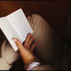May include: A small, white notebook with handwritten text is held open in a person's hand. The notebook is positioned over tan pants. The text is in black ink. The background is a dark brown.