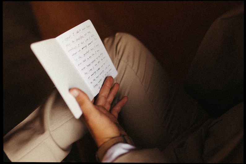 May include: A small, white notebook with handwritten text is held open in a person's hand. The notebook is positioned over tan pants. The text is in black ink. The background is a dark brown.