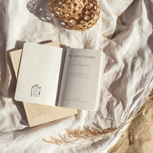 Peut inclure: Une nappe en lin blanc avec un panier tiss&eacute;, un livre intitul&eacute; "The Great Gatsby" de F. Scott Fitzgerald et un carnet avec une illustration dessin&eacute;e &agrave; la main d'un b&acirc;timent. Il y a des citrons sur une assiette blanche et une tige d'herbe s&egrave;che sur le sable.