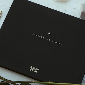 May include: A black book with the words "FOREVER AND ALWAYS" and a star printed on the cover. A gold pen, a gray ring box, and a red rose are also visible. The book has the "Sugar Plum" logo.