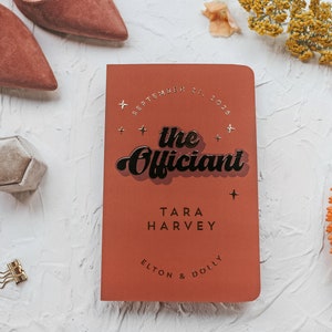 May include: A terracotta-colored wedding officiant book with the text "the Officiant" in a retro font. The book is surrounded by wedding accessories, including shoes, a ring box, and floral accents. The date "September 21, 2025" is also visible.
