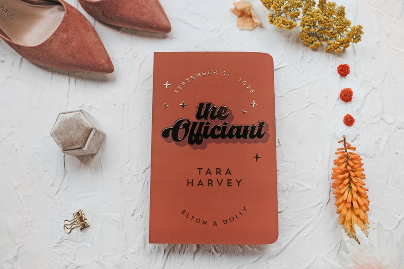 May include: A terracotta-colored wedding officiant book with the text "the Officiant" in a retro font. The book is surrounded by wedding accessories, including shoes, a ring box, and floral accents. The date "September 21, 2025" is also visible.