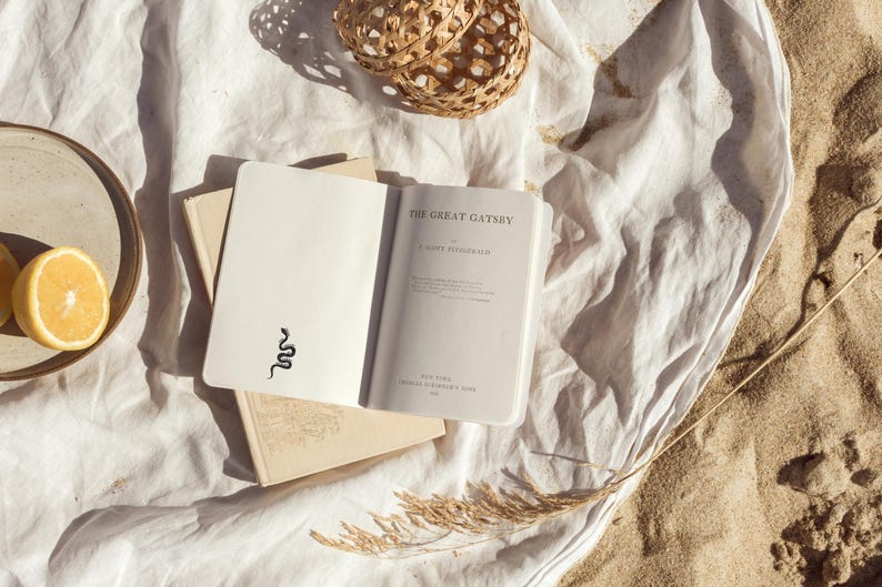 Puede incluir: Un libro abierto titulado "THE GREAT GATSBY" con una ilustraci&oacute;n de serpiente, apoyado en una pila de libros. Tambi&eacute;n se ve una rodaja de lim&oacute;n y una cesta tejida. La escena est&aacute; ambientada sobre una tela blanca y arena.