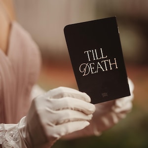 May include: A black card with the words "TILL DEATH" in gold script is held by a person wearing white gloves. The card has a simple, elegant design, suitable for weddings or special occasions.