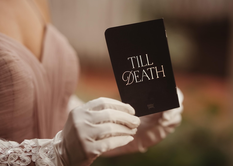 May include: A black card with the words "TILL DEATH" in gold script is held by a person wearing white gloves. The card has a simple, elegant design, suitable for weddings or special occasions.