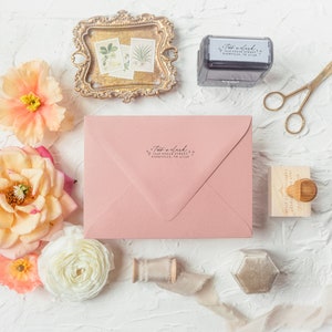 May include: A pink envelope with a return address stamp that reads "Two a clock + 1048 Sugar Street, Huntsville, TN 37756". The envelope is surrounded by flowers, a small wooden stamp, a gold tray with pressed flowers, and a spool of ribbon.