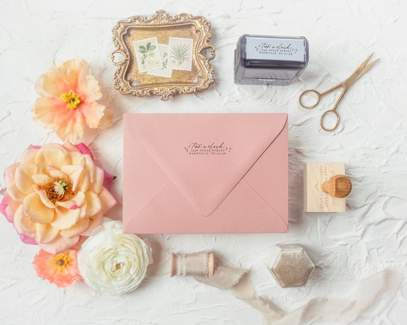 May include: A pink envelope with a return address stamp that reads "Two a clock + 1048 Sugar Street, Huntsville, TN 37756". The envelope is surrounded by flowers, a small wooden stamp, a gold tray with pressed flowers, and a spool of ribbon.
