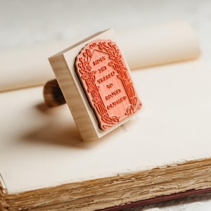 Op de afbeelding: Een houten stempel met een rood rechthoekig ontwerp en de tekst "May your wishes to come true" ligt op een open boek. De stempel heeft een houten handvat en een gedetailleerde bloemenrand. Het boek heeft verouderde, getextureerde pagina's.