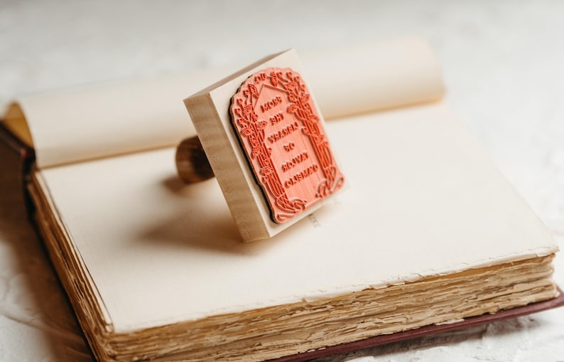 Op de afbeelding: Een houten stempel met een rood rechthoekig ontwerp en de tekst "May your wishes to come true" ligt op een open boek. De stempel heeft een houten handvat en een gedetailleerde bloemenrand. Het boek heeft verouderde, getextureerde pagina's.