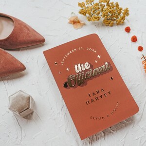 May include: A terracotta-colored notebook with gold lettering that reads "the Officiant" and the date September 21, 2026. A pair of suede, pointed-toe heels and a small, hexagonal ring box are also in the image.