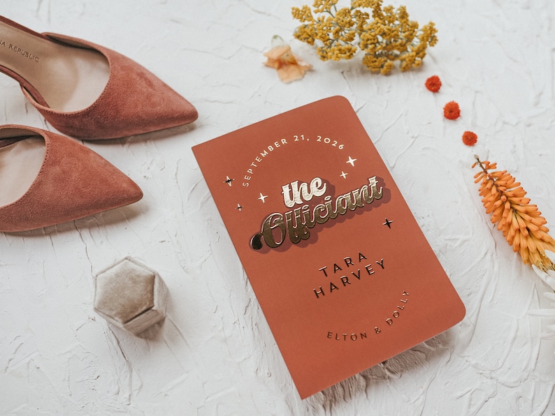 May include: A terracotta-colored notebook with gold lettering that reads "the Officiant" and the date September 21, 2026. A pair of suede, pointed-toe heels and a small, hexagonal ring box are also in the image.