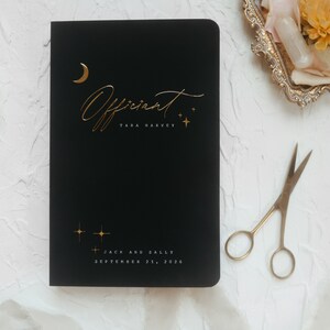 May include: Black notebook with gold foil lettering that reads "Officiant" and the name "Tara Harvey". The notebook also has a gold moon and stars design. The bottom of the notebook reads "Jack and Sally, September 21, 2026".