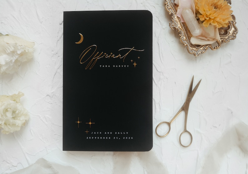 May include: Black notebook with gold foil lettering that reads "Officiant" and the name "Tara Harvey". The notebook also has a gold moon and stars design. The bottom of the notebook reads "Jack and Sally, September 21, 2026".