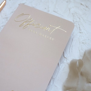 K&ouml;nnte beinhalten: Ein hellrosa Notizbuch mit dem Wort "Officiant" und dem Namen "TARA HARVEY" in goldfarbenen Buchstaben. Ein ros&eacute;goldener Stift und eine Garnrolle sind ebenfalls sichtbar.