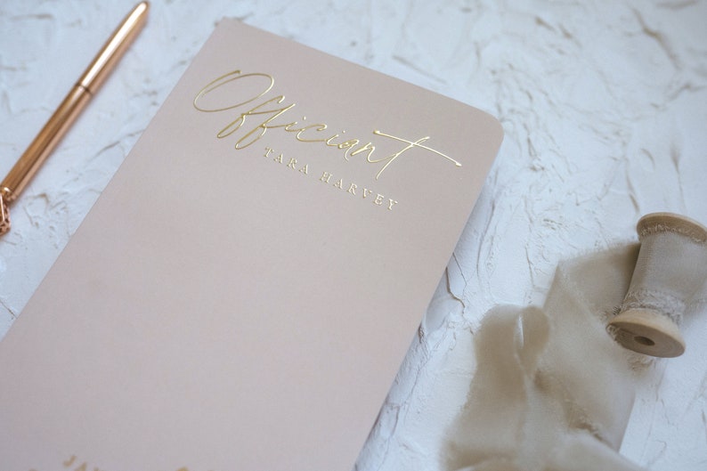 K&ouml;nnte beinhalten: Ein hellrosa Notizbuch mit dem Wort "Officiant" und dem Namen "TARA HARVEY" in goldfarbenen Buchstaben. Ein ros&eacute;goldener Stift und eine Garnrolle sind ebenfalls sichtbar.