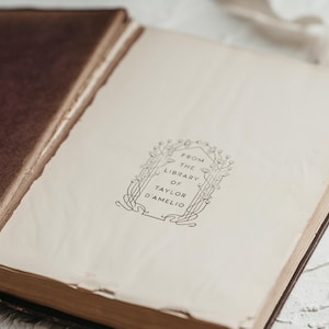 Op de afbeelding: Een open antiek boek met verouderde, cr&egrave;mekleurige pagina's. De kaft van het boek is diepbruin en de pagina's tonen de tekst "FROM THE LIBRARY OF TAYLOR D'AMELIO" in een sierlijke lijst. Het boek is licht geopend.