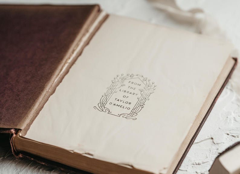 Op de afbeelding: Een open antiek boek met verouderde, cr&egrave;mekleurige pagina's. De kaft van het boek is diepbruin en de pagina's tonen de tekst "FROM THE LIBRARY OF TAYLOR D'AMELIO" in een sierlijke lijst. Het boek is licht geopend.