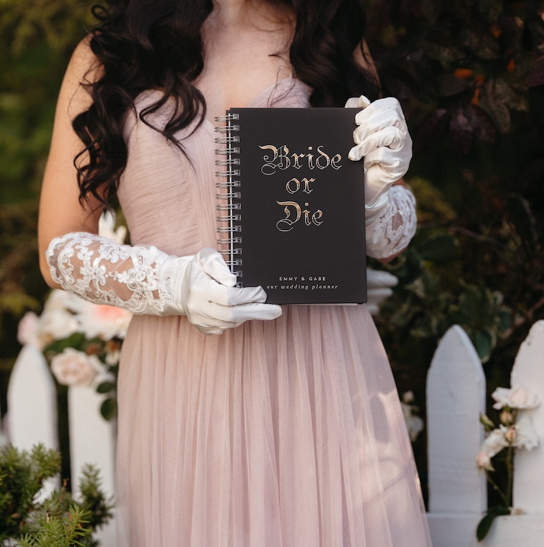 May include: A black spiral-bound wedding planner with the words "Bride or Die" in gold script. The planner also includes the text "Emmy & Gabe our wedding planner". The planner is held by a person wearing white gloves and a pink dress.