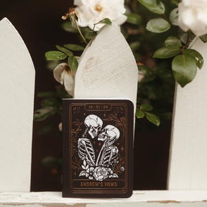 May include: A dark brown notebook with a gold border and the text "10-31-20" at the top. The cover features a black and white illustration of two skeletons kissing, with the words "Andrew's Vows" at the bottom.