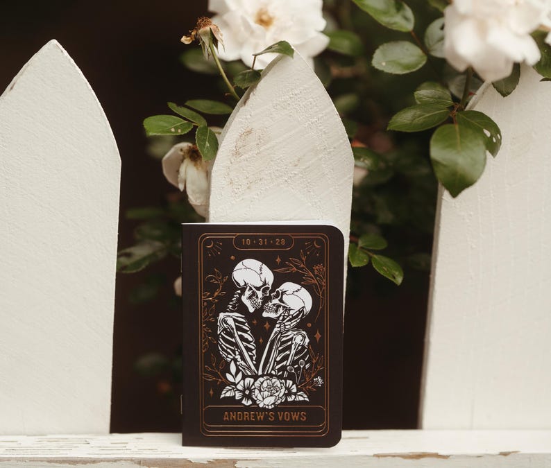 May include: A dark brown notebook with a gold border and the text "10-31-20" at the top. The cover features a black and white illustration of two skeletons kissing, with the words "Andrew's Vows" at the bottom.