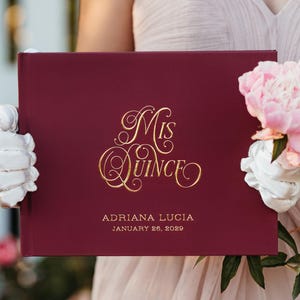 May include: A rectangular burgundy photo album with gold foil lettering that reads Mis Quince, Adriana Lucia, and January 26, 2029. The cover features a smooth matte finish and elegant script typography for the title, centred above the name and date.