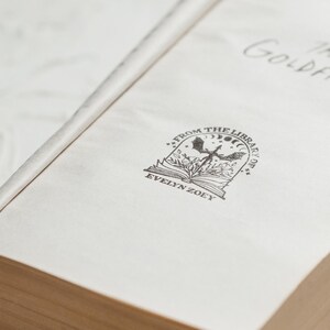 May include: The cover of a book titled "The Goldfinch" with a bookplate that reads "From the Library of Evelyn Zoey" and features a black and white illustration of a bird with wings spread, a book, and a crescent moon.