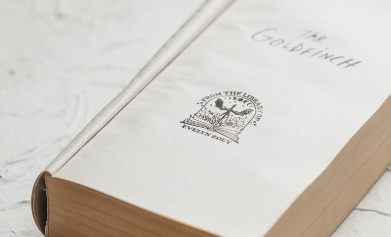 May include: The cover of a book titled "The Goldfinch" with a bookplate that reads "From the Library of Evelyn Zoey" and features a black and white illustration of a bird with wings spread, a book, and a crescent moon.