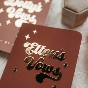 May include: Two burgundy red vow books with gold foil lettering. The top book says "Dolly's Vows" and the bottom book says "Elton's Vows".