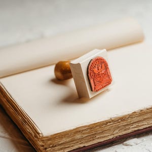 May include: A wooden stamp with a red rubber design of a bird and a book. The stamp is on top of an open book with blank pages.