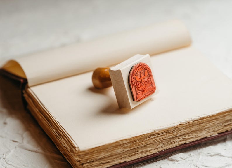 May include: A wooden stamp with a red rubber design of a bird and a book. The stamp is on top of an open book with blank pages.