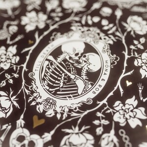 以下が含まれることがあります： 黒と白の装飾的なプリントで、花のフレームの中で抱き合う骸骨が描かれています。「Till Death」の文字と「Mona & Rob」の名前が見えます。 金色のハートのアクセントと追加の骸骨の姿がデザインを完成させています。