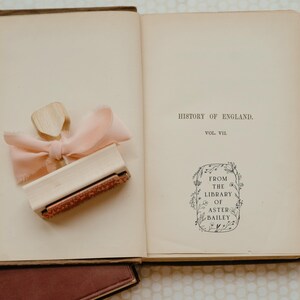 May include: An open book with a brown leather cover. The book is titled "History of England, Vol. VII". A wooden stamp with a pink ribbon is on the left page. The stamp has a floral design and the text "From the Library of Aster Bailey".