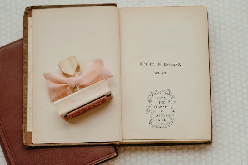 May include: An open book with a brown leather cover. The book is titled "History of England, Vol. VII". A wooden stamp with a pink ribbon is on the left page. The stamp has a floral design and the text "From the Library of Aster Bailey".