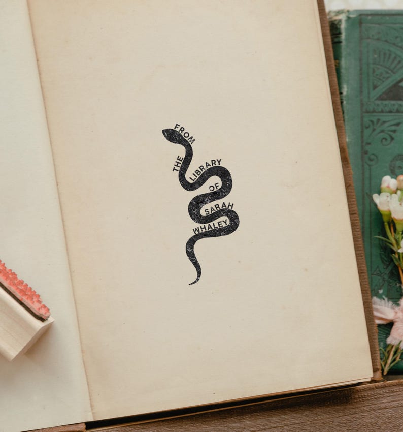 Puede incluir: P&aacute;gina de un libro antiguo con un sello negro en forma de serpiente que dice "FROM THE LIBRARY OF SARAH WHALEY". El sello est&aacute; centrado en la p&aacute;gina. Un sello de madera es visible en el lado izquierdo. El libro est&aacute; abierto.