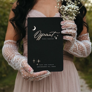 May include: A black card with gold lettering that reads "Officiant Tara Harvey" and "Jack and Sally September 21, 2026". The card has a crescent moon and stars in the top left corner and bottom right corner.