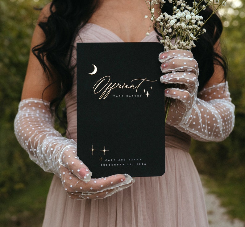 May include: A black notebook with gold lettering that reads "Officiant" and the name "Tara Harvey". The notebook also includes the names "Jack and Sally" and the date "September 21, 2026". The notebook is held by a person wearing white gloves.