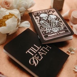 May include: Two black notebooks with gold lettering and accents. One notebook reads "Till Death" and the other reads "Frankie's Vows" with a skeleton couple illustration. The notebooks are surrounded by flowers and ribbon.