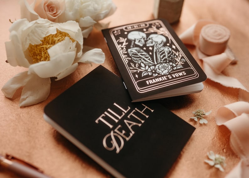 May include: Two black notebooks with gold lettering and accents. One notebook reads "Till Death" and the other reads "Frankie's Vows" with a skeleton couple illustration. The notebooks are surrounded by flowers and ribbon.