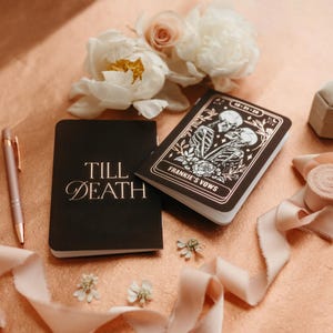 May include: A black notebook with the words "Till Death" in gold script sits next to a pen and a decorative notebook with the words "Frankie's Vows". The notebooks are on a peach-colored surface with flowers and ribbon.