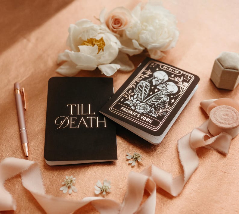 May include: A black notebook with the words "Till Death" in gold script sits next to a pen and a decorative notebook with the words "Frankie's Vows". The notebooks are on a peach-colored surface with flowers and ribbon.