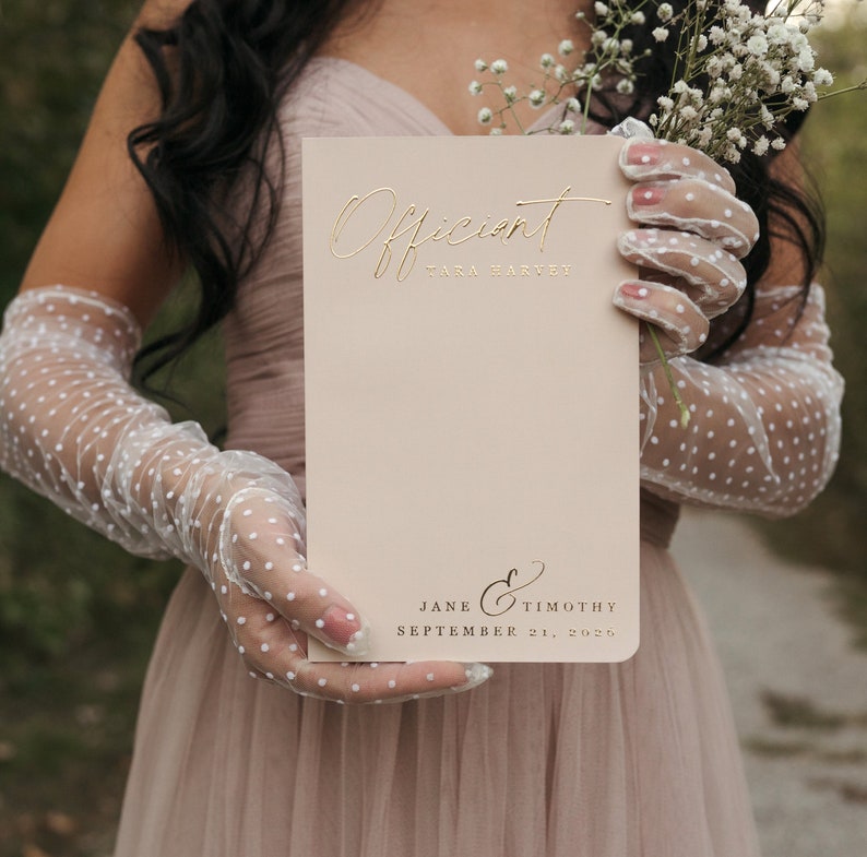 K&ouml;nnte beinhalten: Eine hellbeige Hochzeitskartenkarte mit goldfarbenem Schriftzug "Officiant" und "Tara Harvey". Auf der Unterseite der Karte stehen die Namen "Jane & Timothy" und das Datum "21. September 2025".