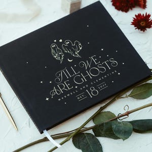 May include: Black hardcover book with the text "Till We Are Ghosts" and the date Oct. 18, 2021. The cover features illustrations of two ghosts and stars. A gold pen and red roses are also in the image.