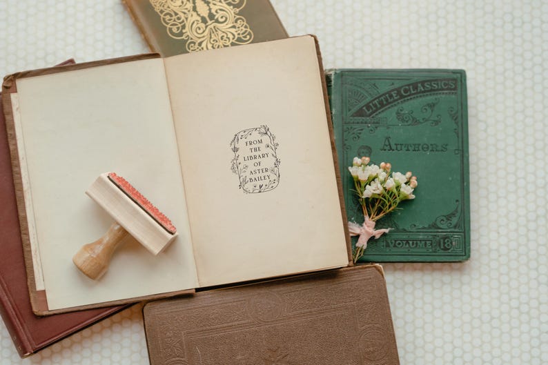 May include: A close-up of an open book with a brown cover. The book has a stamp that reads "From the Library of Aster Bailey". There is a wooden stamp and a small green book with the text "Little Classics Authors Volume 181" on the cover.