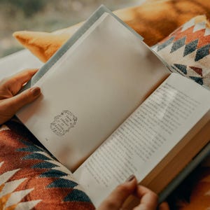 May include: A person is reading a book with a white cover and a decorative design on the front. The book is open and the pages are filled with text. The person is sitting on a colorful blanket with a geometric pattern.