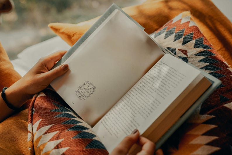May include: A person is reading a book with a white cover and a decorative design on the front. The book is open and the pages are filled with text. The person is sitting on a colorful blanket with a geometric pattern.